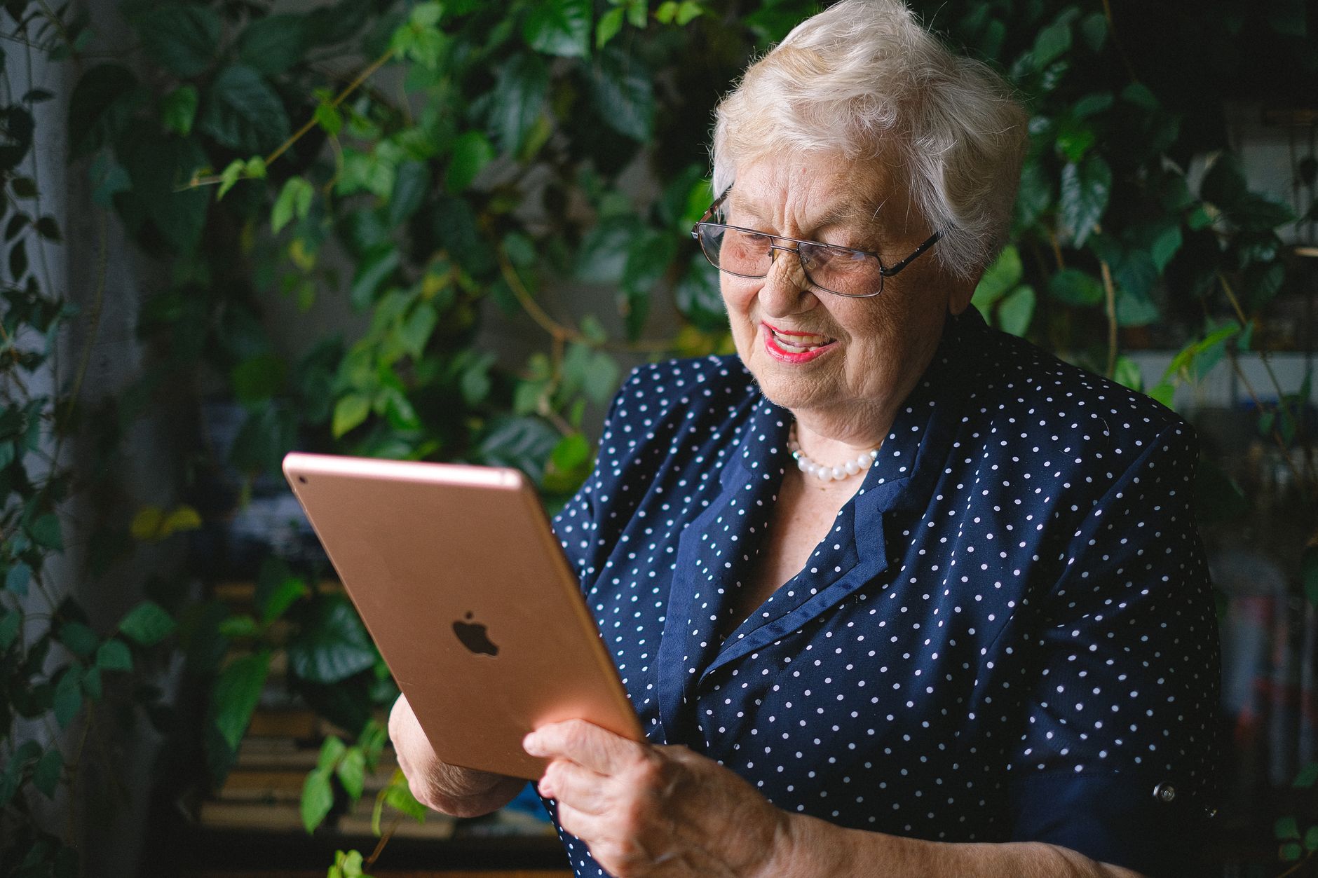 Smiling woman in glasses and a polka-dot top holding a tablet in a room full of green plants